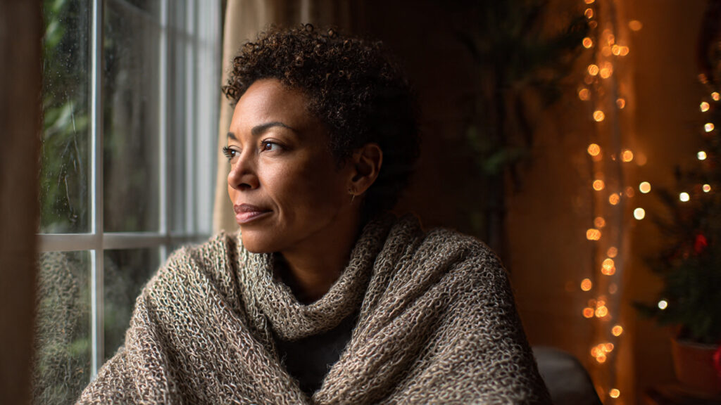 Black woman in her 40s sitting by a window during the holidays, looking thoughtful and calm in soft, warm light.