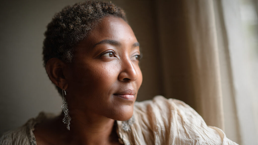 Black woman in her mid-40s sitting by a window, looking thoughtful and reflective, representing midlife disappointment and self-awareness